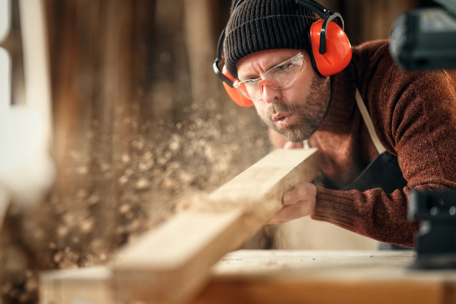 Tischler mit Gehörschutz bläst Späne von einer frisch gehobelten Bohle